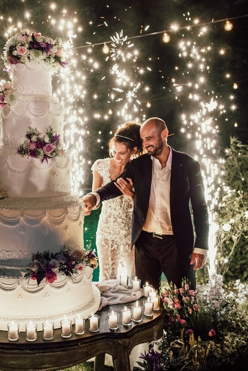 Bride and groom cut large wedding cake with fireworks behind them
