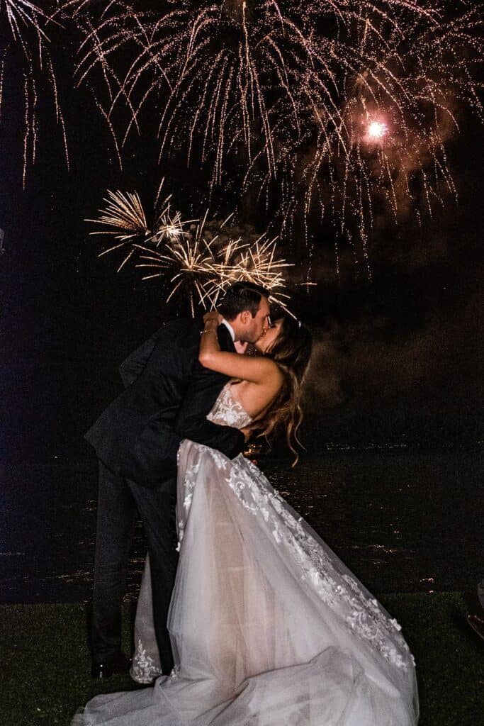 Bride and groom kiss with fireworks in background at wedding reception, taken by photographer who focuses solely on weddings after learning how to outsource for photographers